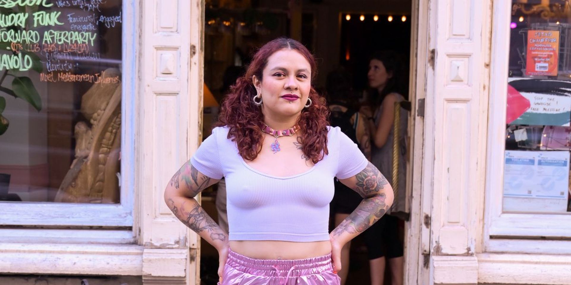 A woman with curly hair and tattoos stands confidently in front of a shop, wearing a light purple crop top and shiny pink shorts.