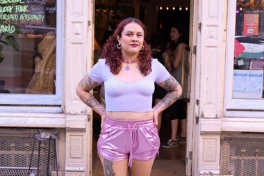 A woman with curly hair and tattoos stands confidently in front of a shop, wearing a light purple crop top and shiny pink shorts.