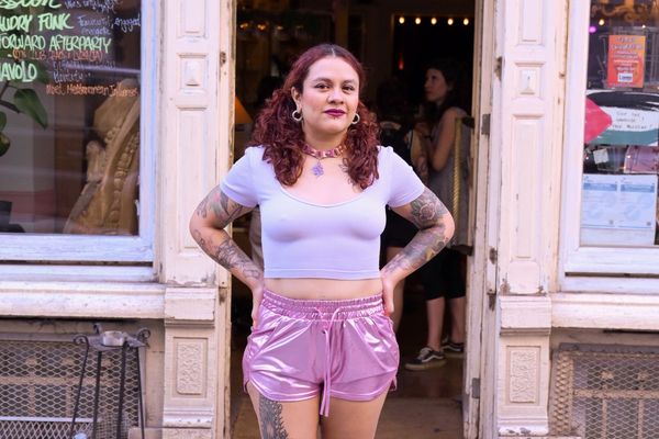 A woman with curly hair and tattoos stands confidently in front of a shop, wearing a light purple crop top and shiny pink shorts.