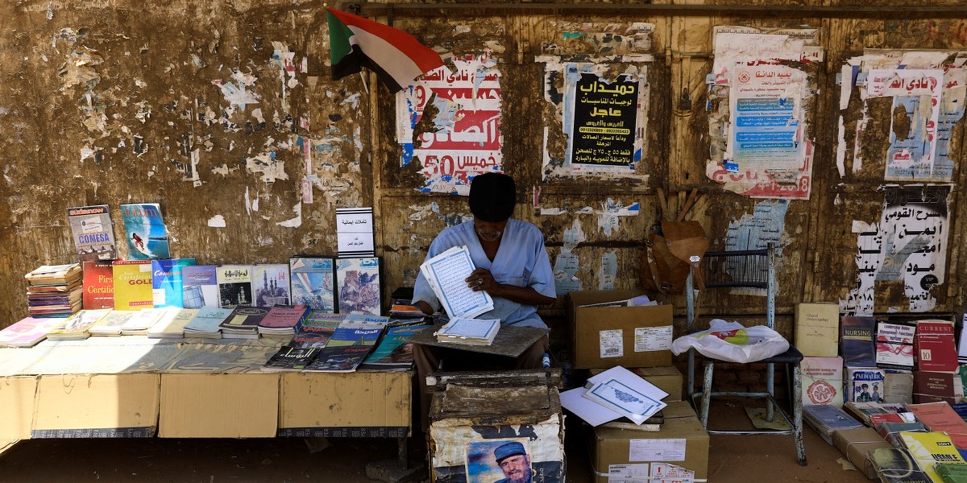 A street vendor in front of a wall of posters sells a variety of books beneath a small Sudanese flag, with boxes and a chair nearby.