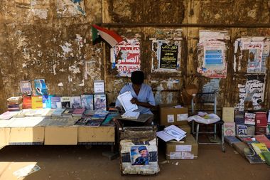 A street vendor in front of a wall of posters sells a variety of books beneath a small Sudanese flag, with boxes and a chair nearby.