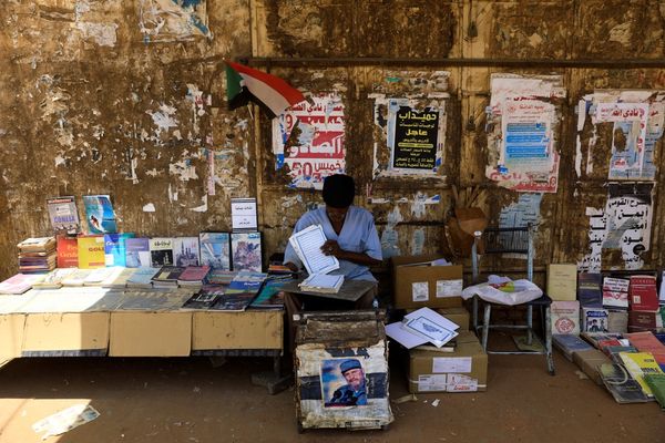 A street vendor in front of a wall of posters sells a variety of books beneath a small Sudanese flag, with boxes and a chair nearby.