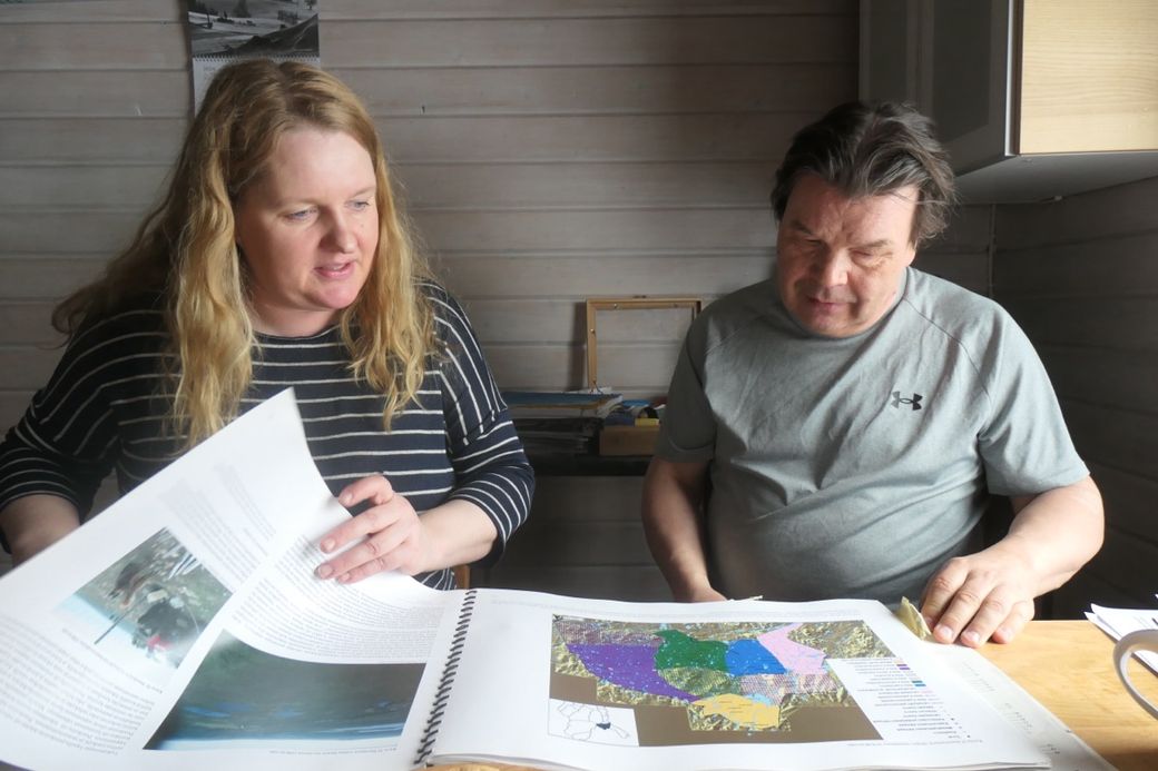 Two people sit at a table reviewing a document with colorful maps, inside a wood-paneled room.