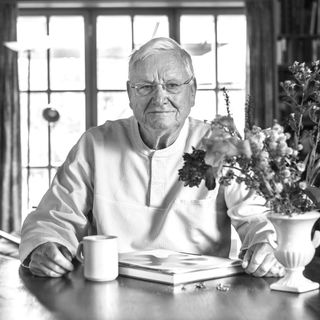 Elderly man in white shirt sitting at a table with a book, mug, and vase of flowers, in a warmly lit room with bookshelves and windows.