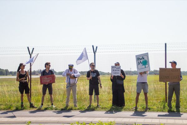 A group of protesters stand by a fence in a field, holding signs and flags with messages advocating for park openings and clean air.