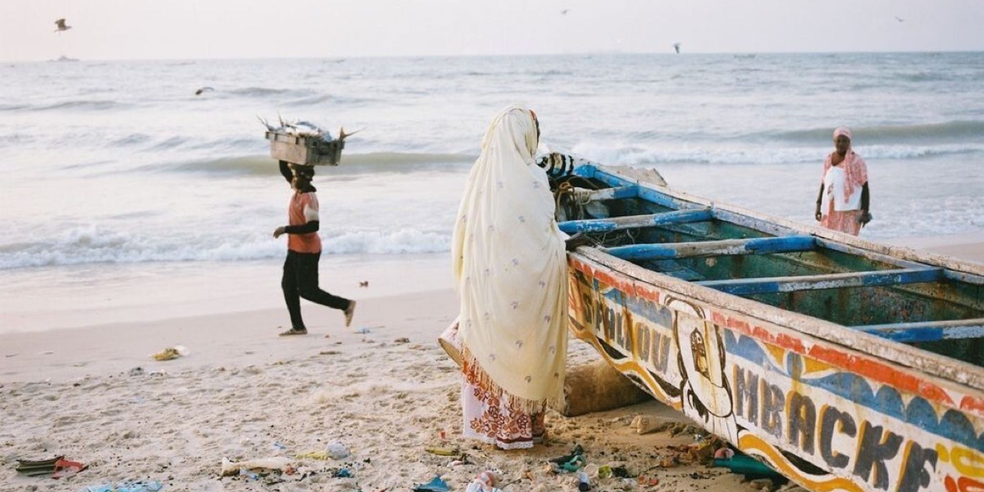 Person in a shawl leans on a colorful boat on a littered beach. Another person carries a crate of fish on their head. Seagulls fly over the sea.