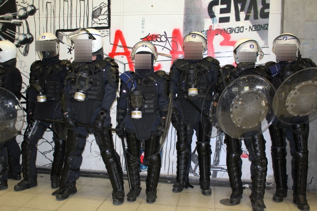 A line of police officers in riot gear, holding shields and batons, stand in front of graffiti-covered walls indoors.