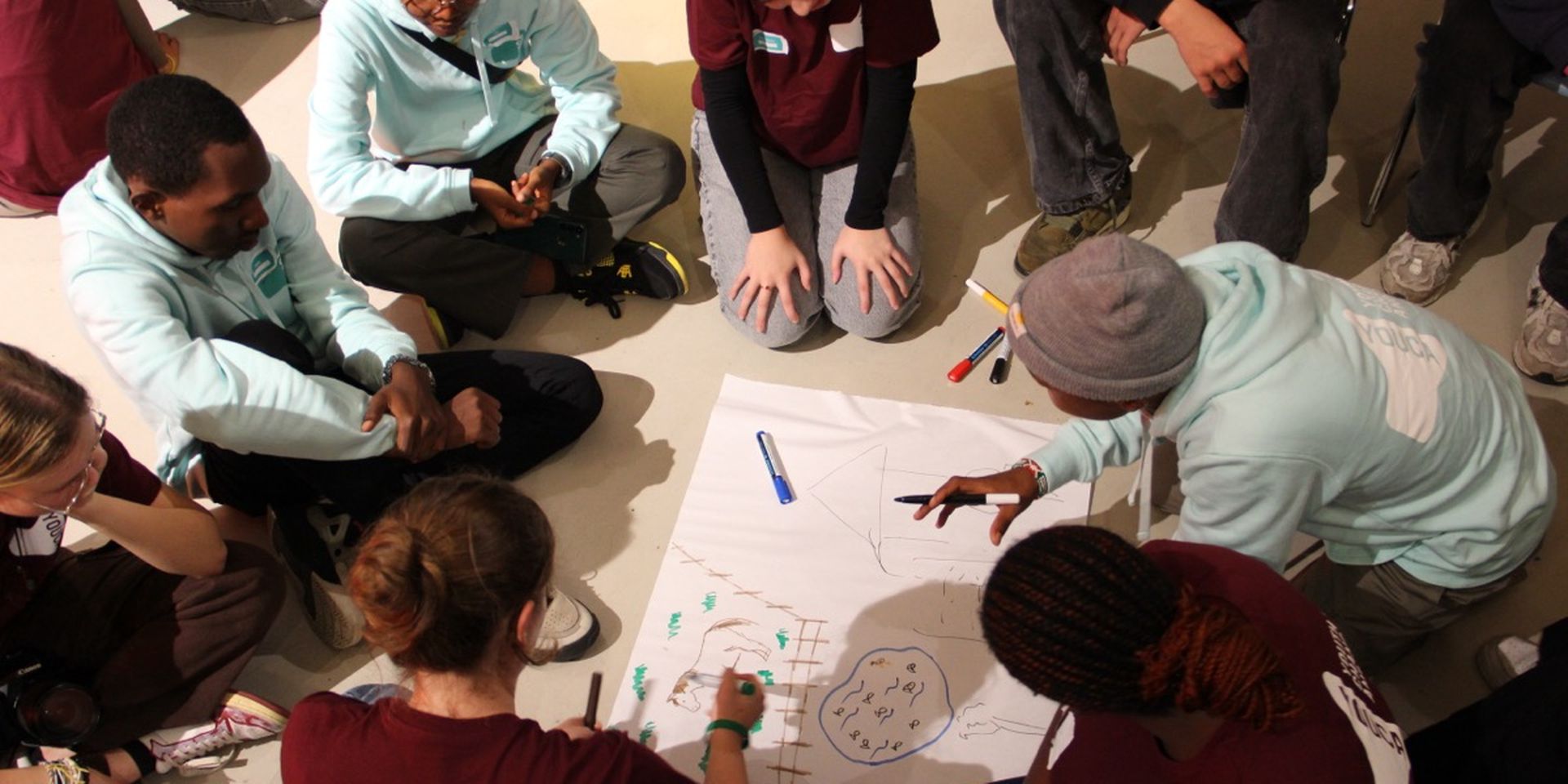 A group of young people sitting in a circle, collaborating on a drawing on large paper, with markers scattered around.