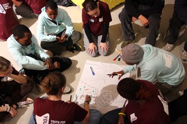 A group of young people sitting in a circle, collaborating on a drawing on large paper, with markers scattered around.