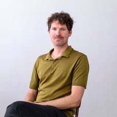 A man with curly hair in a green shirt sits casually against a plain background, looking at the camera.