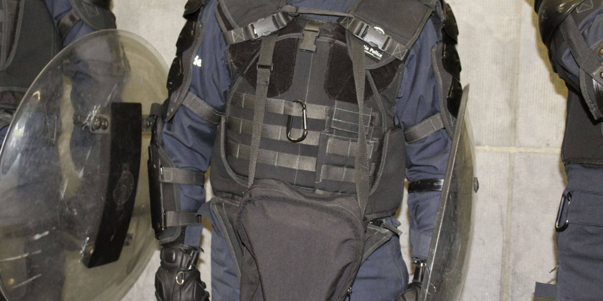 Riot gear-clad police officer with protective vest, gloves, and shield, standing against a concrete wall indoors.