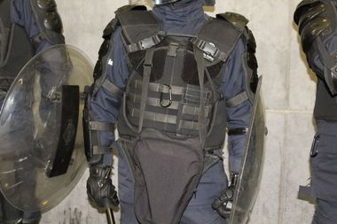Riot gear-clad police officer with protective vest, gloves, and shield, standing against a concrete wall indoors.