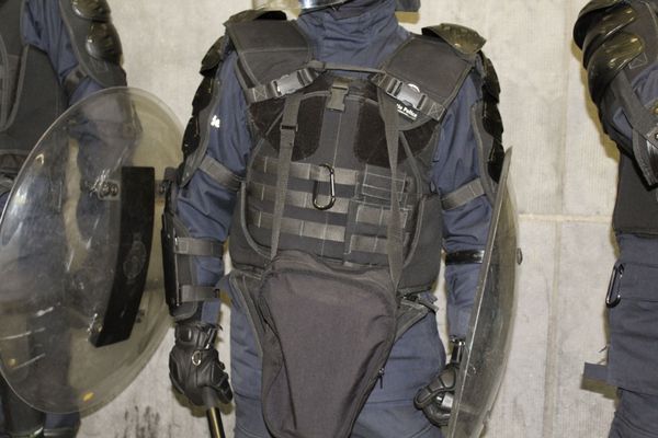 Riot gear-clad police officer with protective vest, gloves, and shield, standing against a concrete wall indoors.