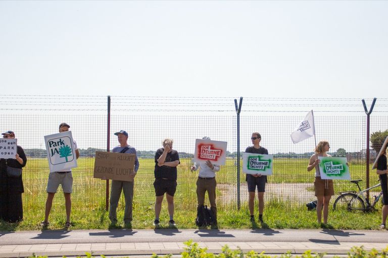 Protesters stand near a fence holding signs and banners advocating for environmental changes and closing Deurne, on a sunny day.