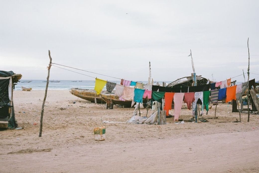 Colorful clothes hanging on a line at a sandy beach, with old boats and the ocean in the background.