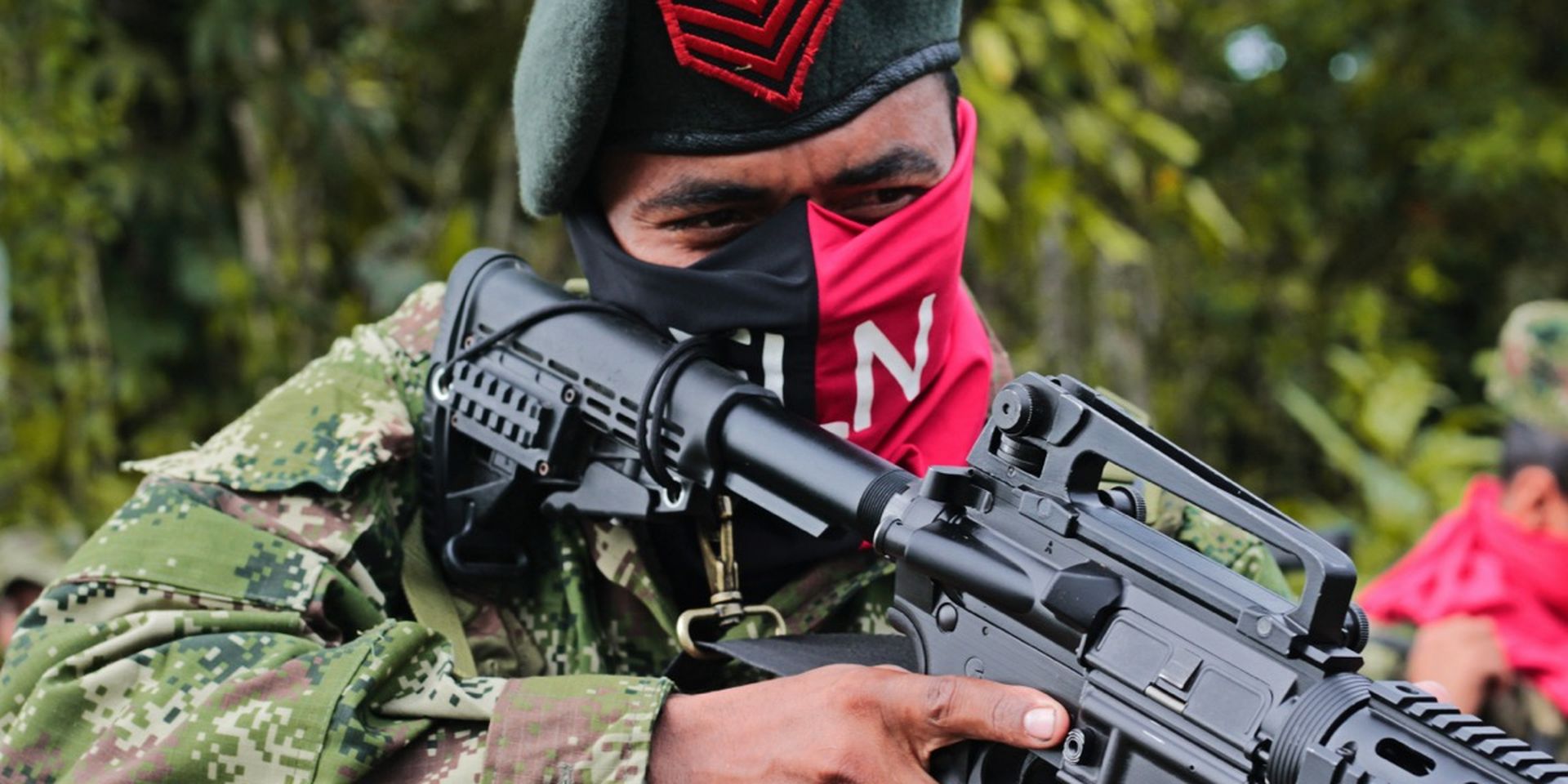 Armed individual in camouflage holds rifle, face partially covered, wearing a beret with a red emblem, standing in a forest setting.