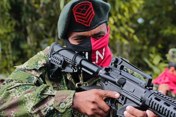 Armed individual in camouflage holds rifle, face partially covered, wearing a beret with a red emblem, standing in a forest setting.