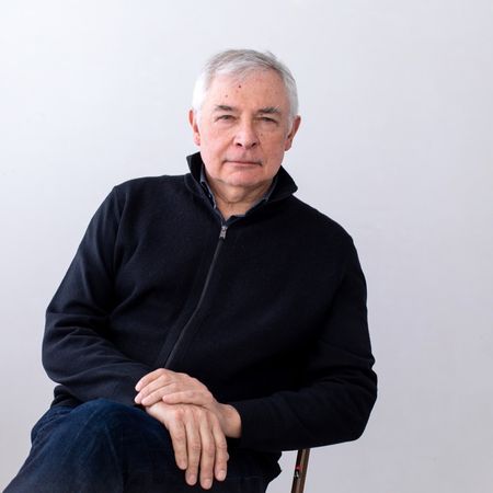 Older man with gray hair seated, wearing a black zip-up jacket against a plain white background.