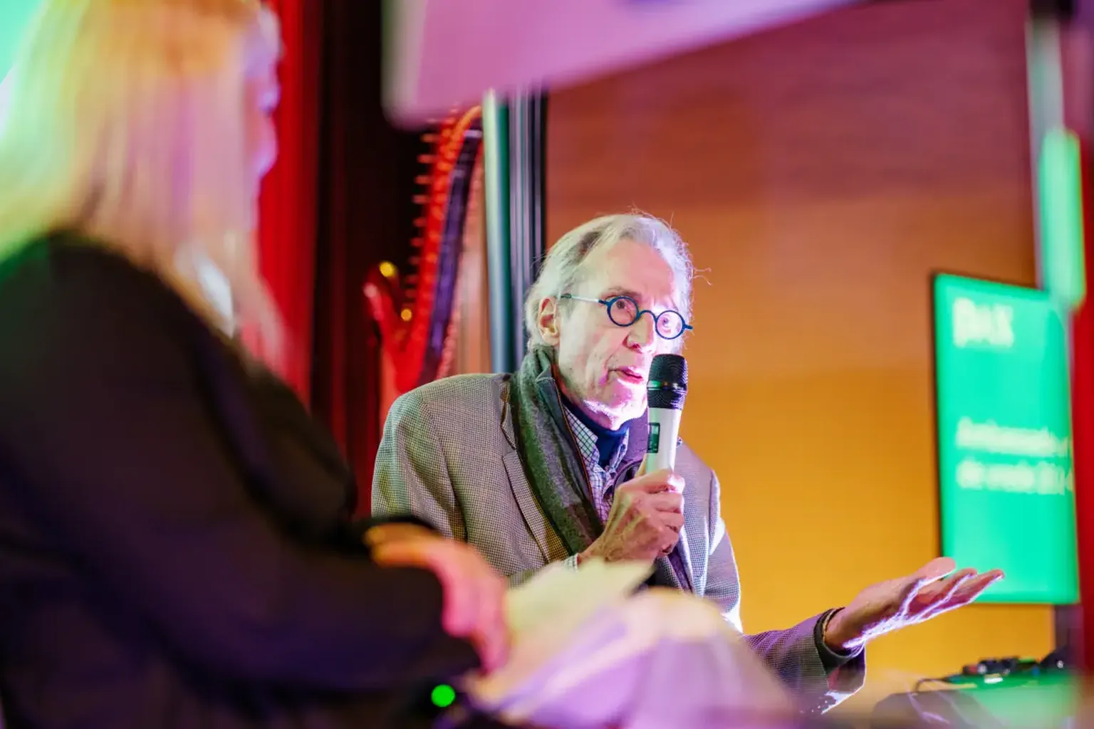 An older man speaks into a microphone during an event, seated beside a woman with long blonde hair, under colorful lighting.