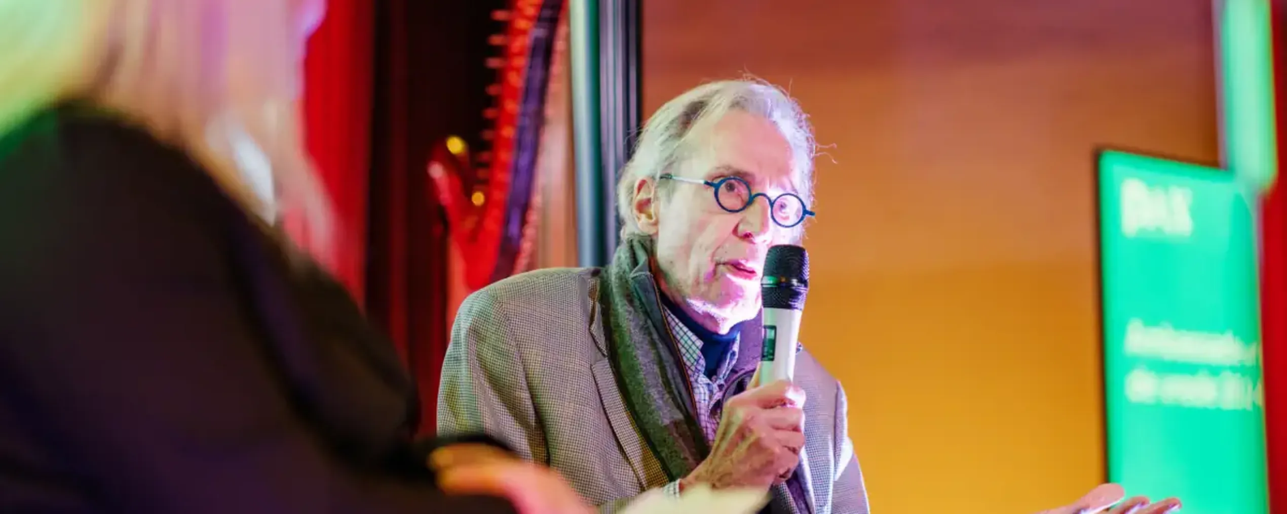 An older man speaks into a microphone during an event, seated beside a woman with long blonde hair, under colorful lighting.