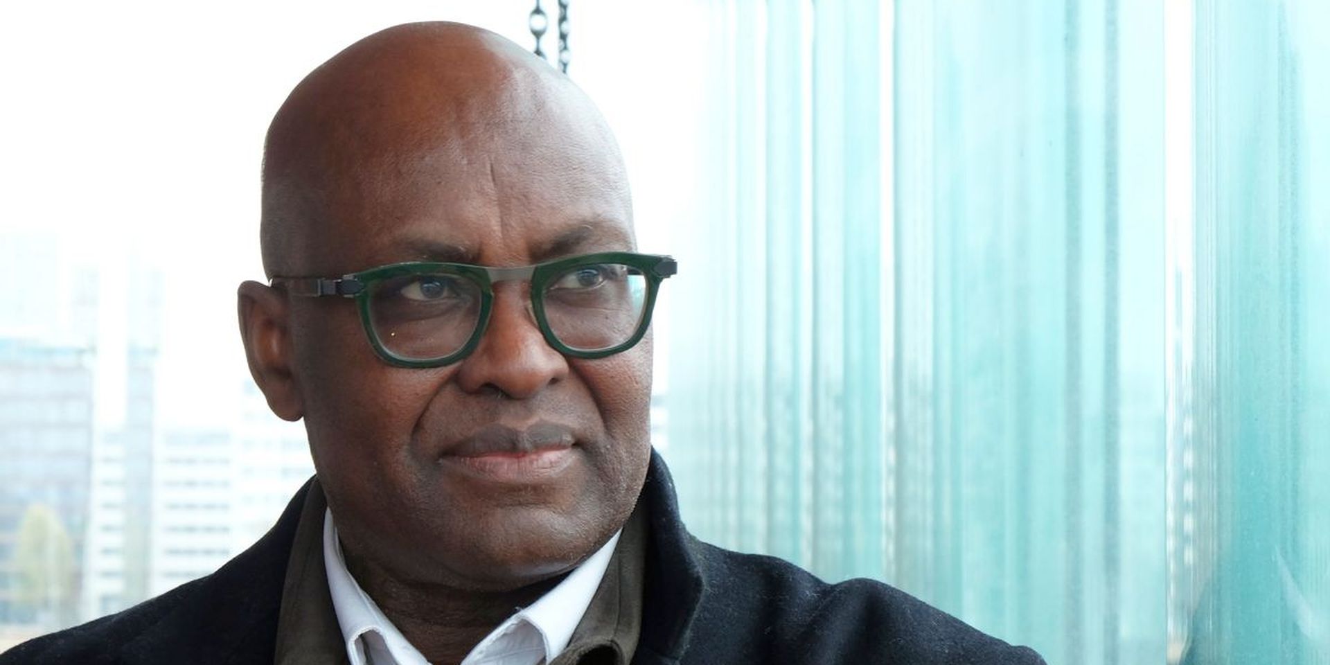 Achille Mbembe kijkt bedachtzaam. Op de achtergrond de ramen van het Antwerpse MAS