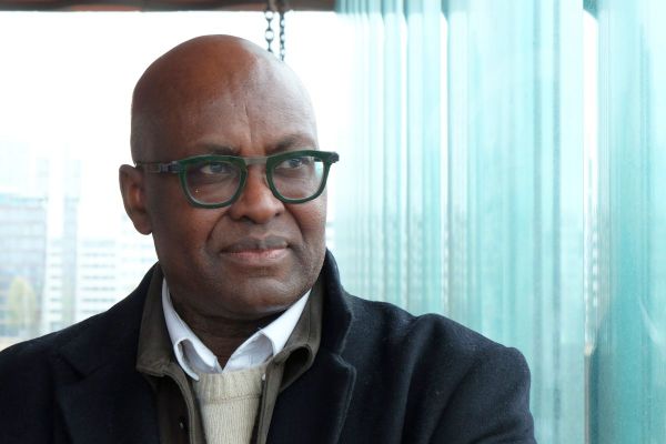 Achille Mbembe kijkt bedachtzaam. Op de achtergrond de ramen van het Antwerpse MAS
