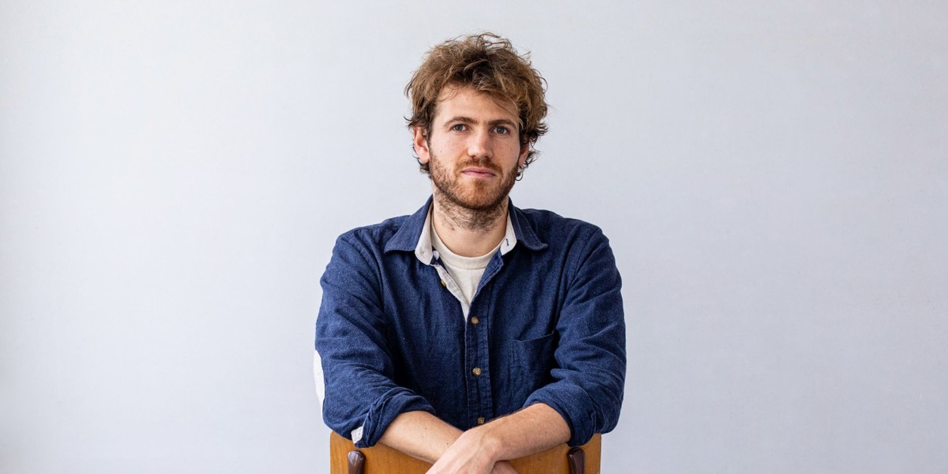 A man with curly hair and a beard leans on a chair, wearing a blue shirt, white t-shirt, and jeans, against a light gray background.