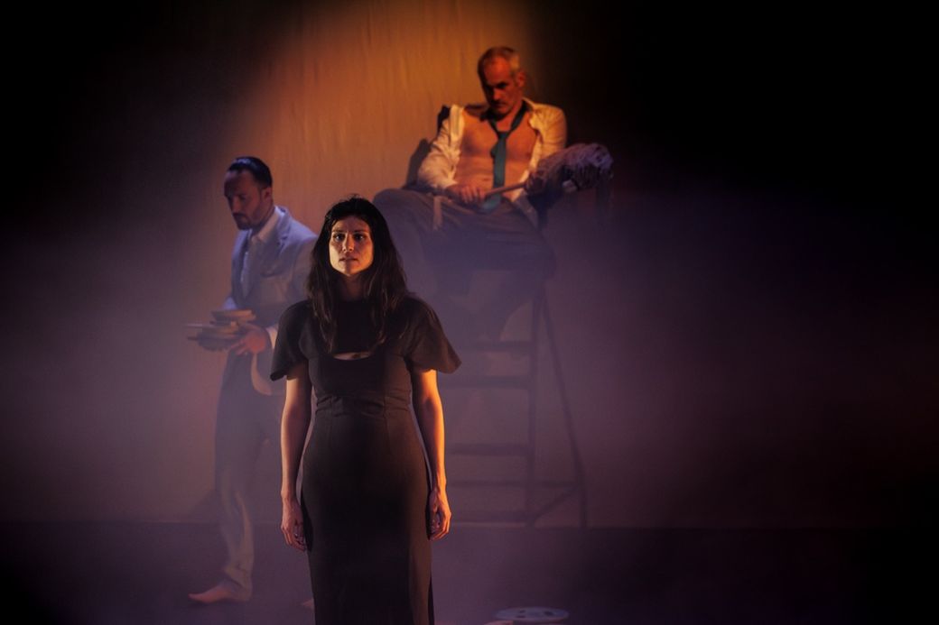 A woman in a dark dress stands in a dimly lit, foggy setting. Two men appear behind her, one seated on a chair, creating a tense atmosphere.