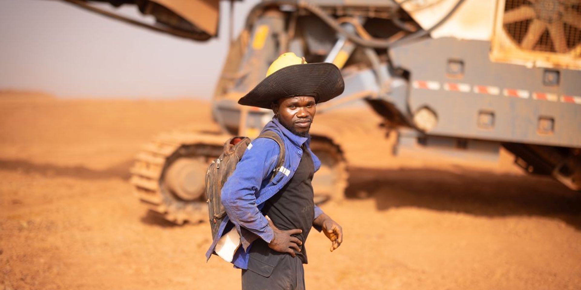 Een man in werkkledij en hoed, met achter hem een grote machine met rupsbanden, op de mijnsite van AG2BAbij Boffa, Guinee.