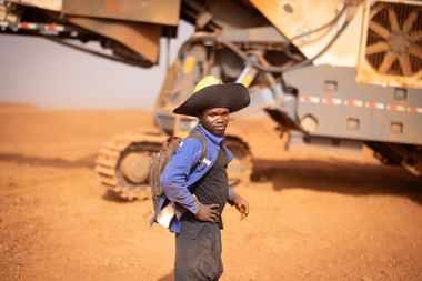 Een man in werkkledij en hoed, met achter hem een grote machine met rupsbanden, op de mijnsite van AG2BAbij Boffa, Guinee.