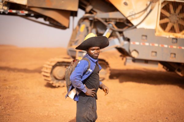 Een man in werkkledij en hoed, met achter hem een grote machine met rupsbanden, op de mijnsite van AG2BAbij Boffa, Guinee.