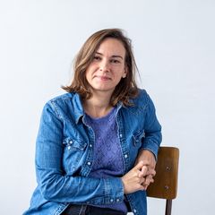 Woman in a denim shirt and purple sweater sits on a chair against a plain white background, smiling gently at the camera.