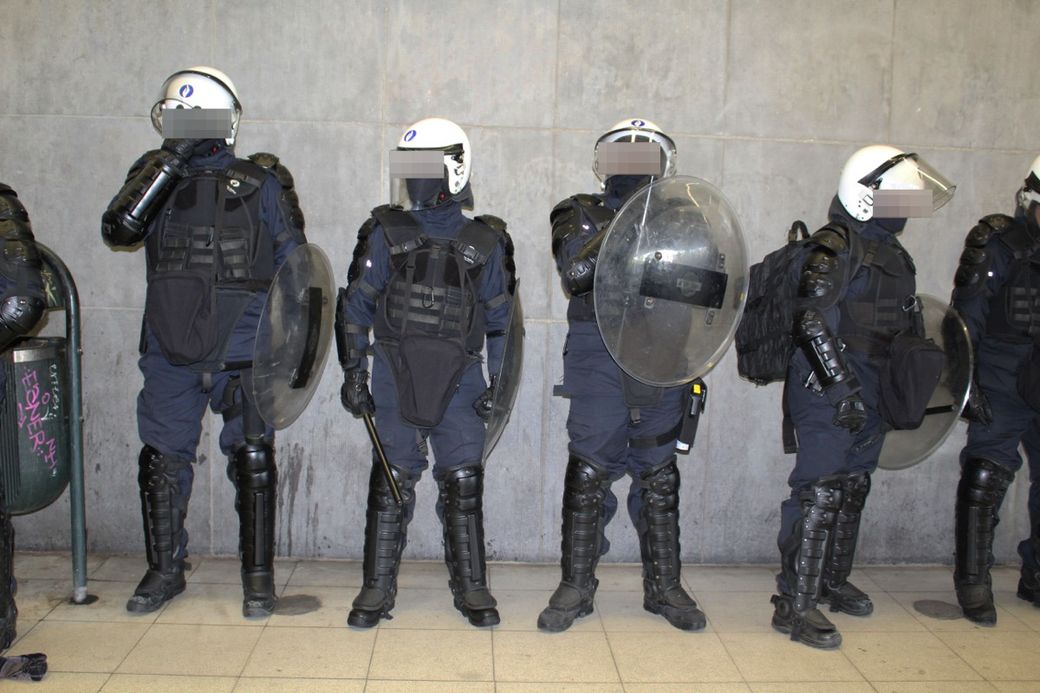 Four riot police officers in protective gear stand against a concrete wall, holding shields and batons.