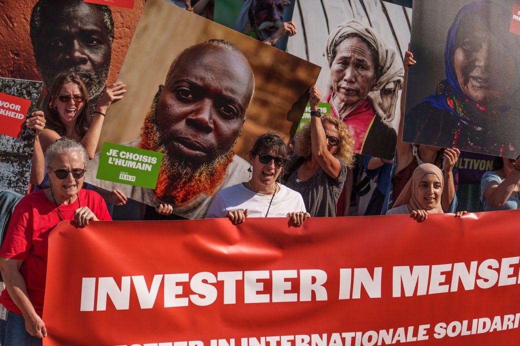A group of people holding a large red banner with the text "INVESTEER IN MENSEN" and large photographs of diverse individuals in the background.