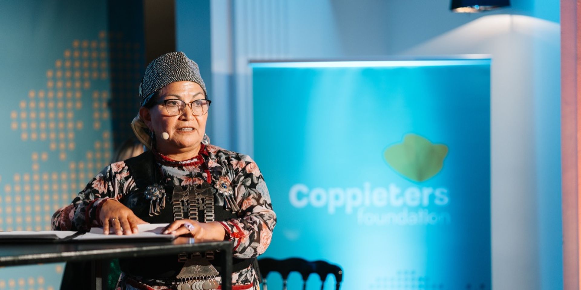 Woman in floral attire speaks at a podium with a microphone. A "Coppieters Foundation" banner is visible in the background.
