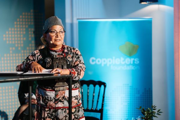 Woman in floral attire speaks at a podium with a microphone. A "Coppieters Foundation" banner is visible in the background.