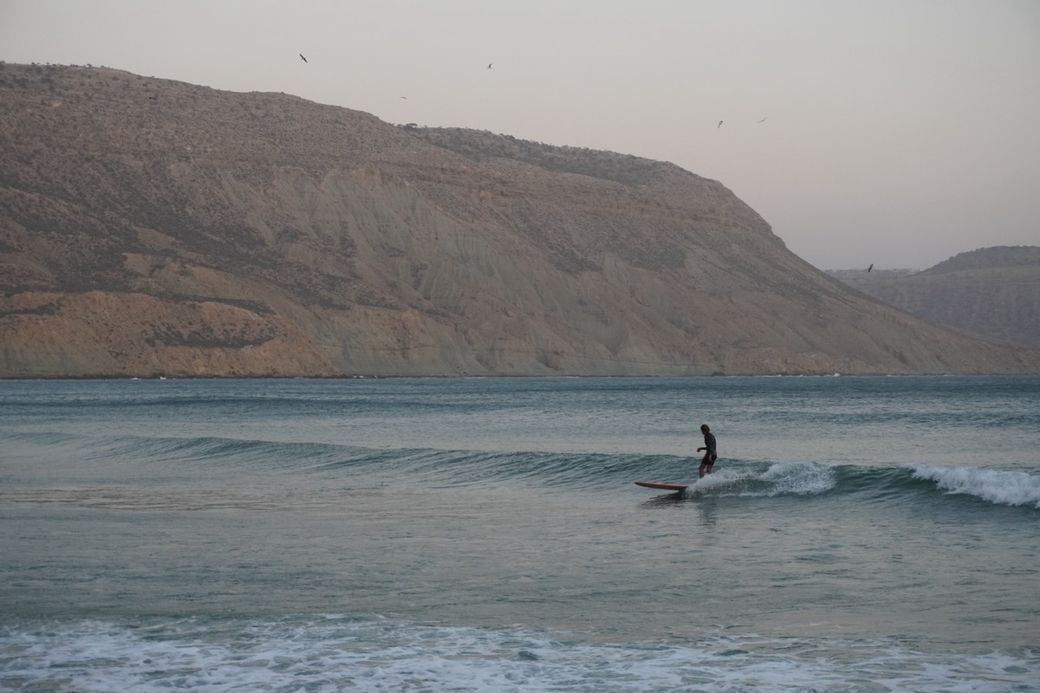 A person surfs a small wave in a calm sea, with a rugged mountainous backdrop and a few birds in the sky.
