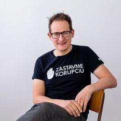 Smiling person with glasses, sitting on a chair, wearing a black t-shirt with "Zastavme Korupciu" printed in white.