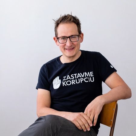 Smiling person with glasses, sitting on a chair, wearing a black t-shirt with "Zastavme Korupciu" printed in white.