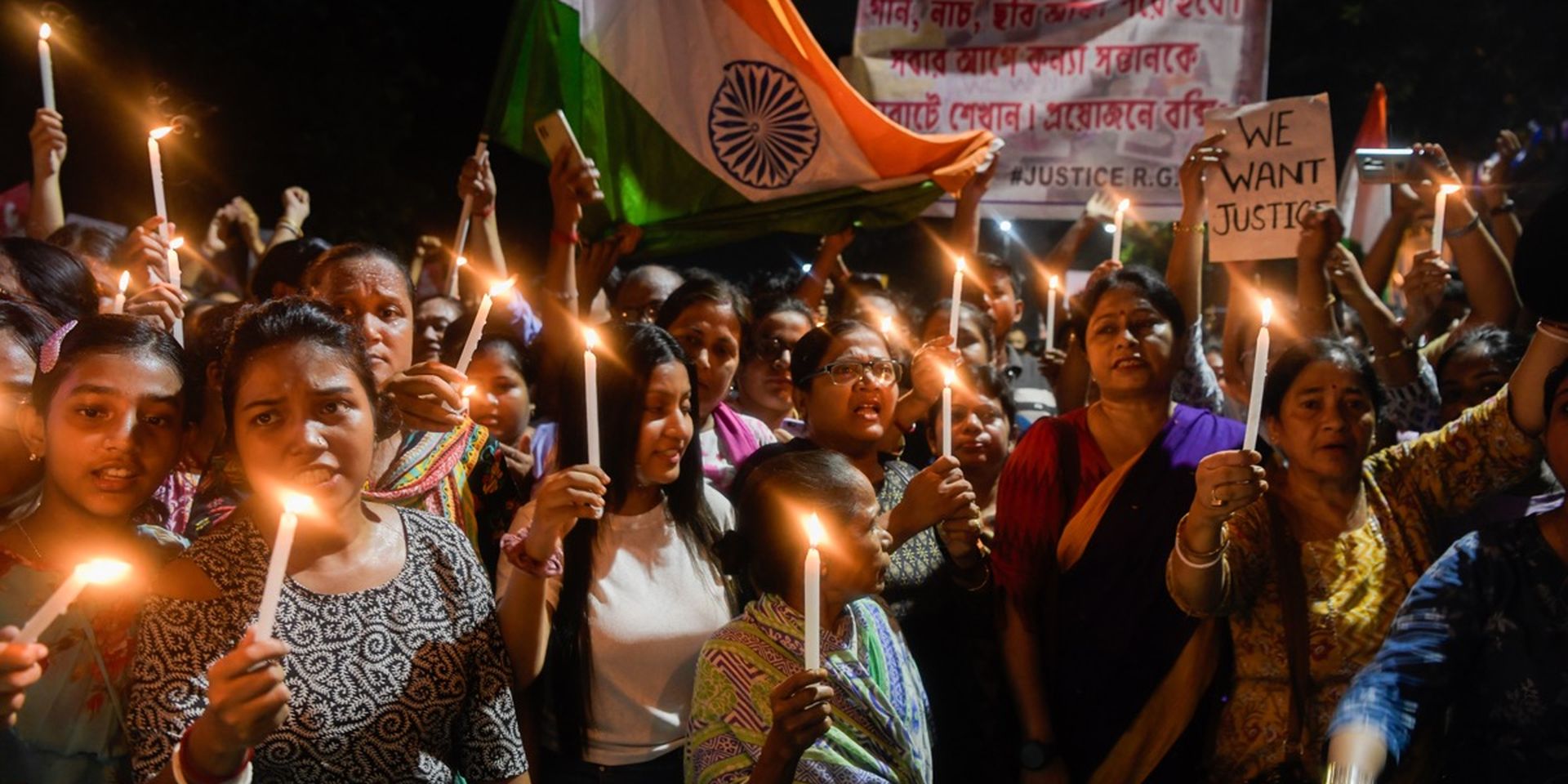 A group of people hold candles during a night protest, displaying signs and an Indian flag, demanding justice.
