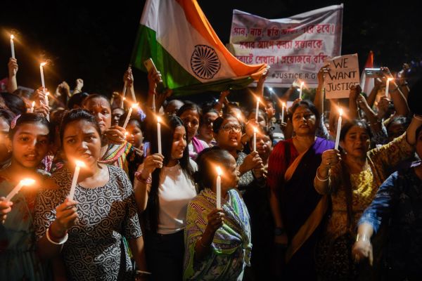 A group of people hold candles during a night protest, displaying signs and an Indian flag, demanding justice.
