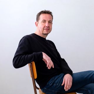 Man in a black sweater and jeans sits casually on a wooden chair against a plain white background.