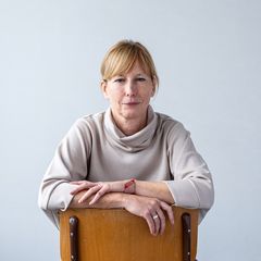 Woman in a light sweater sits backward on a chair, arms resting on the backrest, against a plain background.