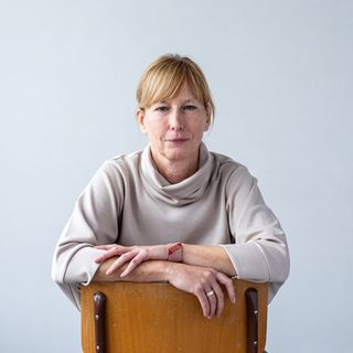 Woman in a light sweater sits backward on a chair, arms resting on the backrest, against a plain background.