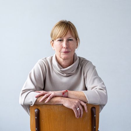 Woman in a light sweater sits backward on a chair, arms resting on the backrest, against a plain background.