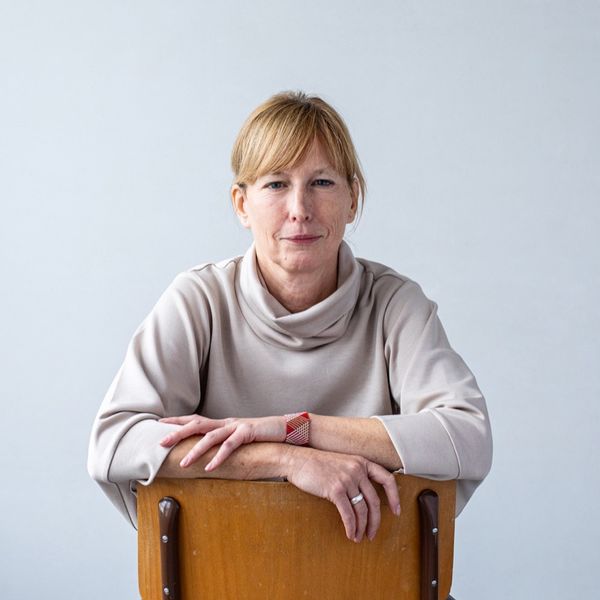 Woman in a light sweater sits backward on a chair, arms resting on the backrest, against a plain background.
