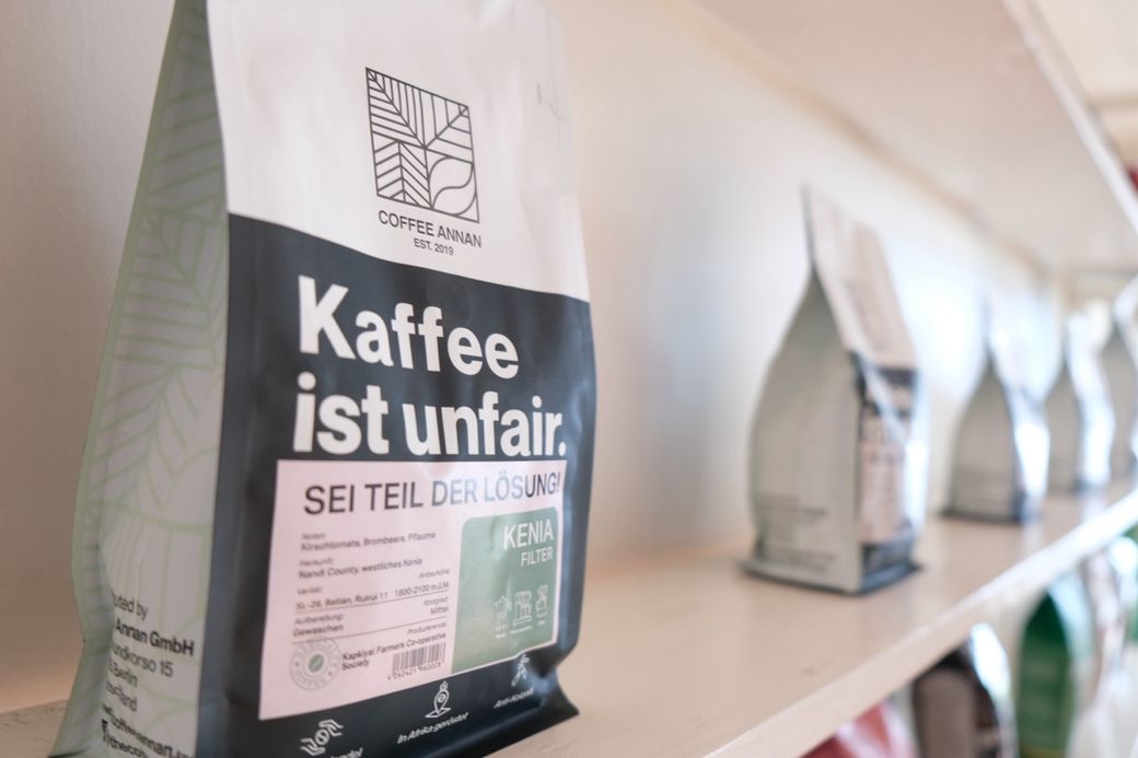 Close-up of a coffee bag labeled "Kaffee ist unfair" on a shelf. "Sei Teil der Lösung" is also visible. Other bags are in the background.