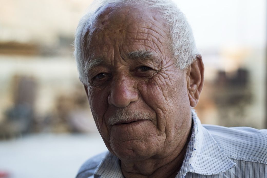 Elderly man with white hair and a lined face, wearing a striped shirt, smiles softly in a softly blurred outdoor setting.