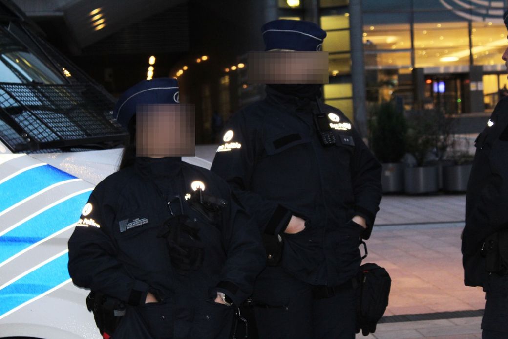 Two female police officers in uniform stand near a police van, one with her hands in her pockets, in an urban setting.