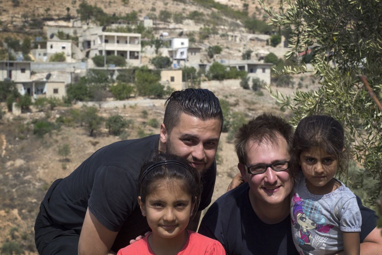 Four people posing outdoors with a hillside village in the background, surrounded by trees and shrubs.